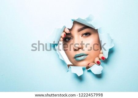 the face of a young beautiful girl with a bright make-up and puffy blue lips peers into a hole in blue paper.Fashion, beauty, make-up, cosmetics, hairstyle, beauty salon, boutique, discounts, sales.