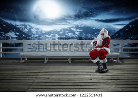Red old Santa Claus and winter night. Wooden pier with white bench and landscape of mountains with lake. Free space for your decoration. Moon light. 