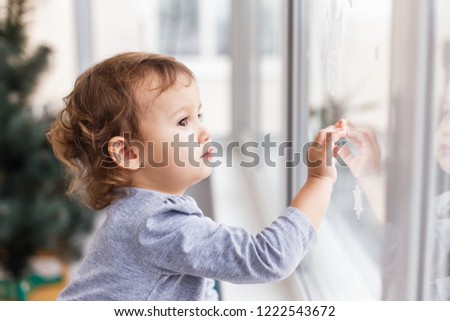 Cute little three years girl looking through window with holiday decoration. Christmas and New Year window decoration
