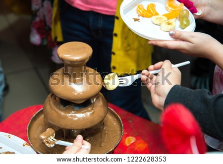 Vibrant Picture of Chocolate  Fontain on children kids birthday party with a kids playing around and marshmallows and fruits dip dipping into fountain.Chocolate fountain placed on a table in w