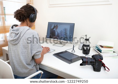 Rear view of young media technology student working on home desk with professional photographic sound equipment, indoors. Black female creative college with picture on screen, lifestyle.