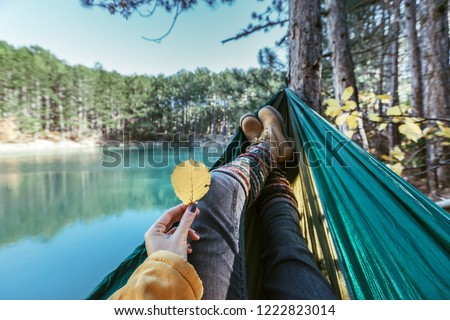 Woman relaxing in the hammock by the lake in the autumn forest, POV view of legs in trekking boots. Hiking in fall. Wanderlust concept scene.