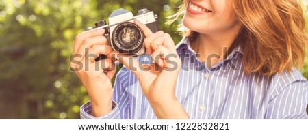Charming smiling young woman in stylish wear taking photos in autumn park using vintage camera, creative professional female photographre making photo during sunny day. Selective focus on camera