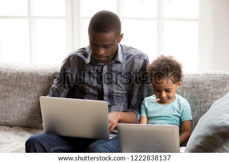 Focused African American family using laptops, computer together at home, serious father and preschooler son sitting on couch near and looking at screen, browsing applications, son repeats after dad