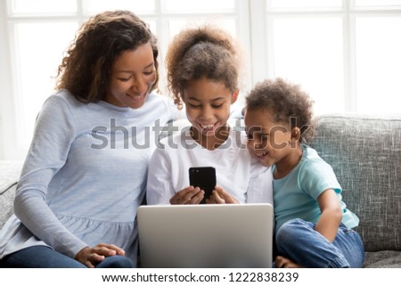Happy African American smiling family using mobile devices together at home, attractive mother and toddler son looking at phone screen, little preschooler daughter holding smartphone and laptop