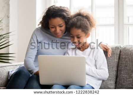 Happy African American attractive mother and little preschooler daughter using laptop, sitting together on couch, sofa in living room at home, watching video, shopping online, looking at screen