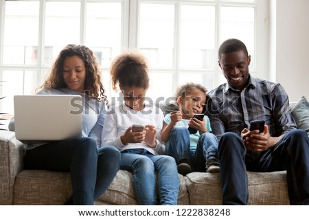 Large African American family using mobile devices, sitting together at home, preschooler daughter, toddler son using phones, mother shopping with laptop, father with smartphone, addicted with devices