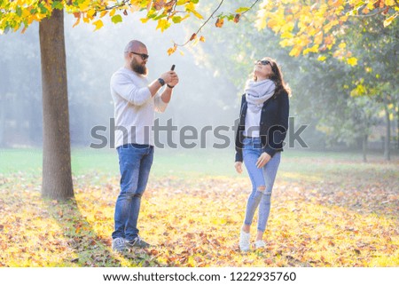 Bearded man photorgaphing his girlfriend with smartphone in sunny autumn garden 