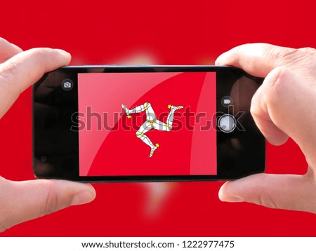 The concept of tourism and travel. The hands of men make a telephone photograph of the flag of Isle Of Man. On the smartphone close-up image of the flag. Photos for social networks, blogs, instagram.