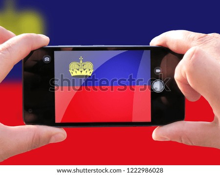 The concept of tourism and travel. The hands of men make a telephone photograph of the flag of Liechtenstein. On the smartphone close-up image of the flag. Photos for social networks, blogs