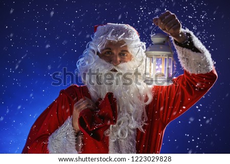 Santa Claus with a long white beard holds candle holder with burning candle against a snowing blue sky. Christmas