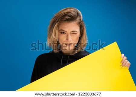Portrait of dark blonde woman winking while holding cardboard paper. Isolated blue background