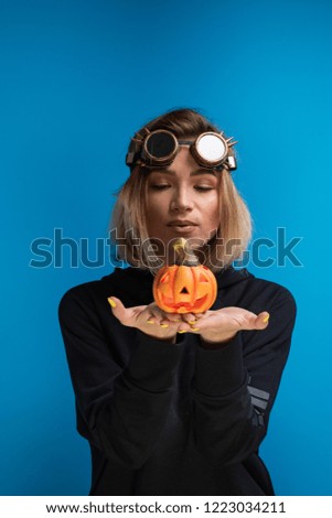 Woman wearing steampunk glasses and black hoodie holding a carved Halloween pumpkin. Themed goth steampunk shooting in the studio against blue background
