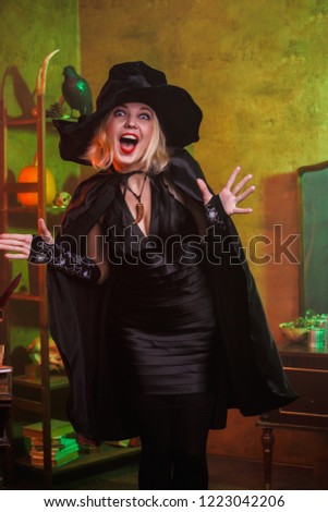 Picture of happy witch in black hat, dress on background of rack with pumpkin and crow