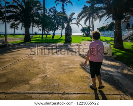 A photo of an old woman enjoying a walk in a beautiful park with palm trees by the Mediterranean Sea. The park is located in the seaside touristic holiday town of Cullera, in Valencia, Spain, Europe.