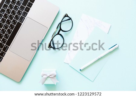 Flatlay of laptop, paper, pen, reading glasses and little gift box on a blue background