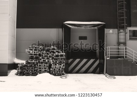 Cargo lift for storage with white black diagonal lines warning sign in grayscale. Wooden pallets on snow. Door for loading goods with copy space. Wall of warehouse in winter. Monochrome building.