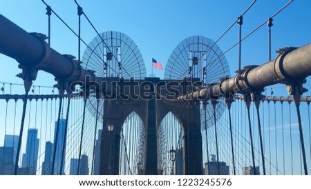 View of Brooklyn Bridge from footpath with pedestrian safety netting and barrier with centralized American Flag aloft bridge tower overlooking Manhattan Skyline