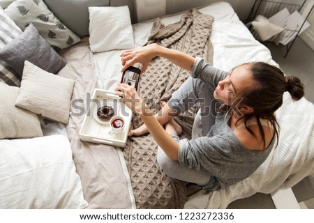From above shot of woman sitting on bed and taking picture of tea and doughnut on tray with smartphone
