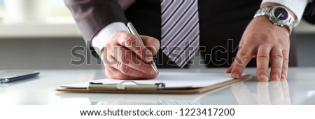 Male arm in suit and tie fill form clipped to pad with silver pen closeup. Sign gesture read pact sale agent bank job make note loan credit mortgage investment finance executive chief legal law