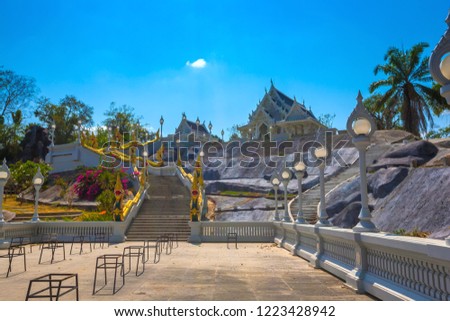 White stone Buddhist temple on a dais, the roof is decorated with carved patterns on wood, a picturesque staircase with golden dragons on the edges. Wat Kaew, Krabi Town, Thailand.