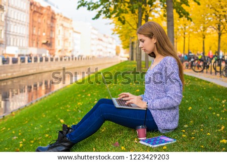 Young woman E-book writer typing article on laptop computer while sitting in autumn park in warm day during recreation time in weekend. Female smart student having online learning via portable netbook