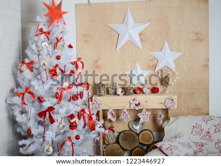 White Christmas tree in the interior, decorated with red bows, balls, toys. Under the Christmas tree are boxes with gifts, decorative stars on the wall and ceiling, and an armchair stands next to it.