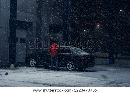Black car parked near office building in winter evening in strong snow storm. Automobile near auto service in night. Driver near machine near automatic gate for maintenance.