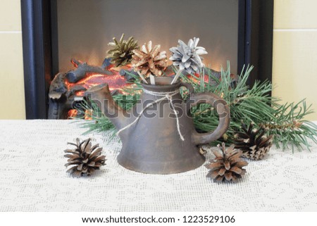 Clay teapot of unusual shape with bouquet of flowers from colorful cones on openwork knitted napkin against the background of pine needles and fireplace.
