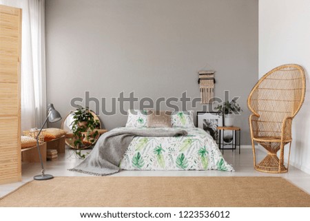 Wicker peacock chair in stylish bedroom interior with natural carpet and king size bed, real photo with copy space and macrame on the empty grey wall