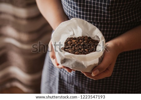 Woman hands holding coffee beans in white paper bag. The woman is wearing a navy blue checkered pinafore. Brown background