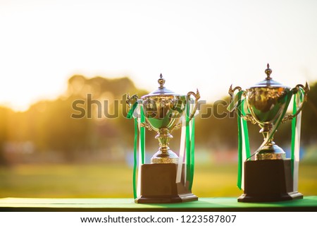 Two silver trophy stand on a green table. The backdrop is a football field. The sun is coming down to reflect the picture becomes golden. Have copy space and  bokeh.