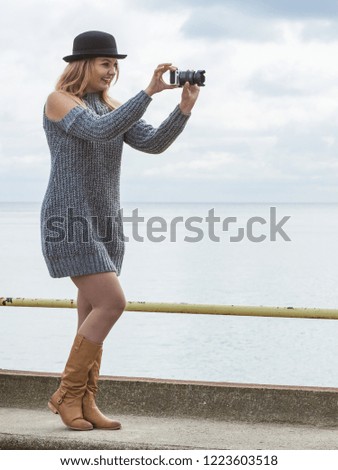 Smiling woman wearing black hat and grey sweater taking pictures using digital camera on outdoor photo session. Female having passion.