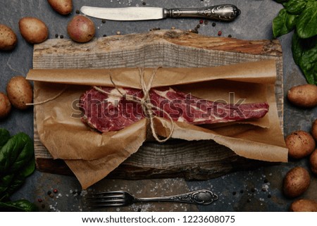 raw rib eye steak wrapped in baking paper on wooden board with spices, potatoes and kitchen cutlery
