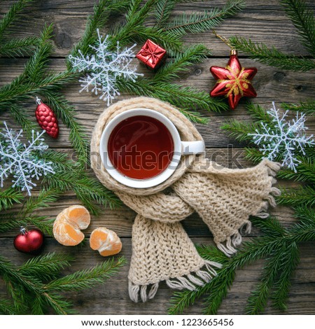 Beautiful cozy Christmas picture with a mug of hot tea in a scarf with fir branches, toys, snowflakes, tangerine on a wooden background