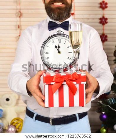 Guy holds striped present box with champagne glass and alarm clock on wooden wall background, defocused. Man with beard holds clock showing almost midnight. Celebration and holiday surprise concept