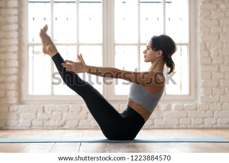 Young sporty attractive woman practicing yoga, doing Paripurna Navasana exercise, balance pose, working out, wearing sportswear, black pants and top, indoor full length, white yoga studio