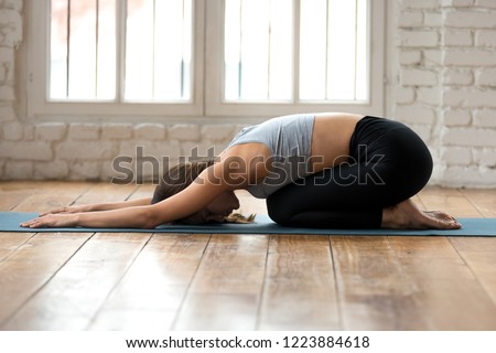 Young sporty attractive woman practicing yoga, doing Child exercise, Balasana pose, working out, wearing sportswear, black pants and top, indoor full length, white yoga studio