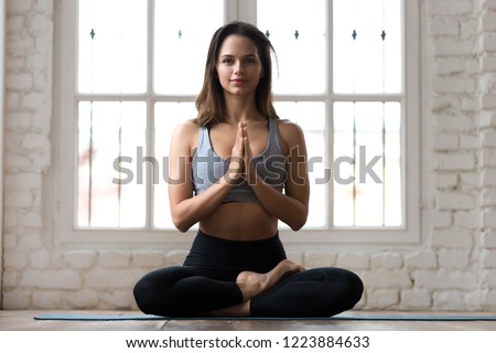 Young smiling attractive sporty woman practicing yoga, doing Ardha Padmasana exercise, meditating in Half Lotus pose with namaste, working out, wearing sportswear, indoor full length, yoga studio Royalty-Free Stock Photo #1223884633
