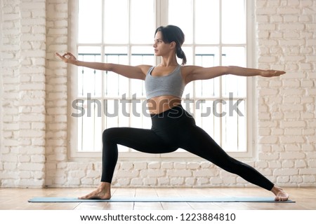 Young sporty attractive woman practicing yoga, doing Warrior Two exercise, Virabhadrasana 2 pose, working out, wearing sportswear, black pants and top, indoor full length, white yoga studio