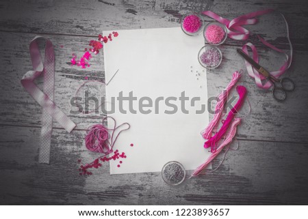 Flat lay with white sheet and pink beads, sequins, floss, scissors and satin ribbons on wooden background top view dramatic toned picture