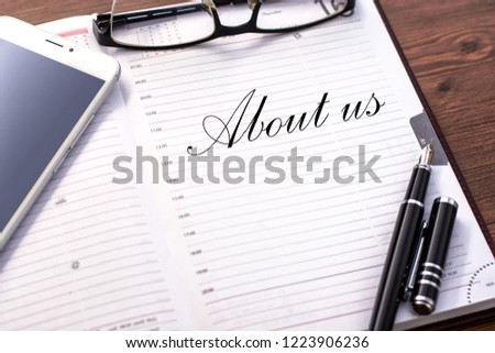 A business notebook (a diary) and  business accessories (mobile phone, glasses, a fountain pen) with text About us.