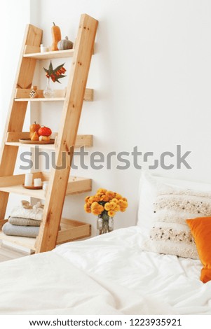 Home decor. Cozy fall bedroom interior: white wall, bed with white linen, light beige plaid, orange pillows, wooden rack, vase with yellow chrysanthemums flowers, pumpkins, candles. Autumn decoration.