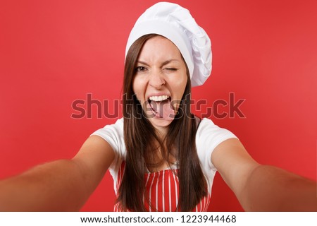 Housewife female chef cook or baker in striped apron, white t-shirt, toque chefs hat isolated on red wall background. Close up housekeeper woman doing taking selfie shot. Mock up copy space concept