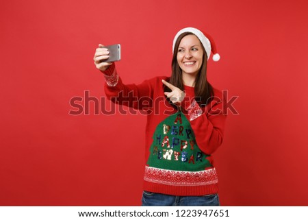 Smiling young Santa girl doing taking selfie shot on mobile phone, pointing index finger isolated on bright red background. Happy New Year 2019 celebration holiday party concept. Mock up copy space