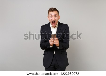 Excited young business man keeping mouth open wide, looking surprised using mobile phone typing sms message isolated on grey background. Achievement career wealth business concept. Mock up copy space