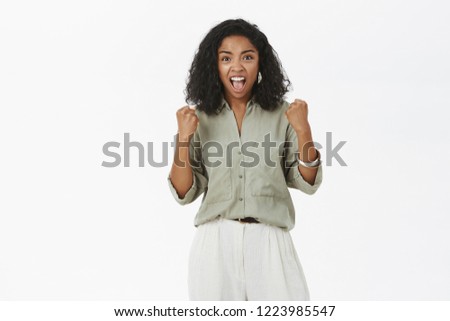 Studio shot of excited supportive energized dark-skinned woman with curly hairstyle raising clenched fists while cheering for favorite team yelling joyfully posing against gray background