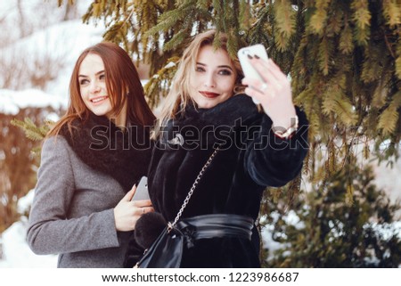 two fashionable ladies standing in a winter park near tree and use the phone