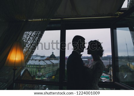 beautiful and elegant blonde bride in a long white dress with her handsome bride in a blue suit standing in the hotel room near window