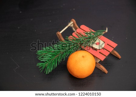 Tangerines with leaves in a tiny toy sledge, surrounded by Christmas decor with Christmas tree, dry orange and candies over wooden back ground, sledge orange tree